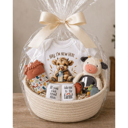 Baby gift basket with a cow onesie, cow mirror plush toy, cow teething mitt, and farm socks in clear cellophane wrap and rope basket on a wooden table top.