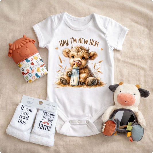 Baby cow 4pc gift set with onesie "Hay I'm new here highland cow image", cow teething mitt, cow plush mirror toy, and take me to the farm baby socks on a creme background.