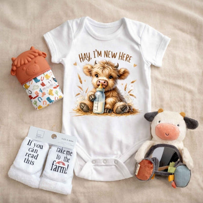 White onesie with little cow drinking bottle and says "Hay I'm new Here" on front on a tan fabric background with a cow mitten teether, plush mirror cow toy, and farm theme baby socks. Little Gift Nook