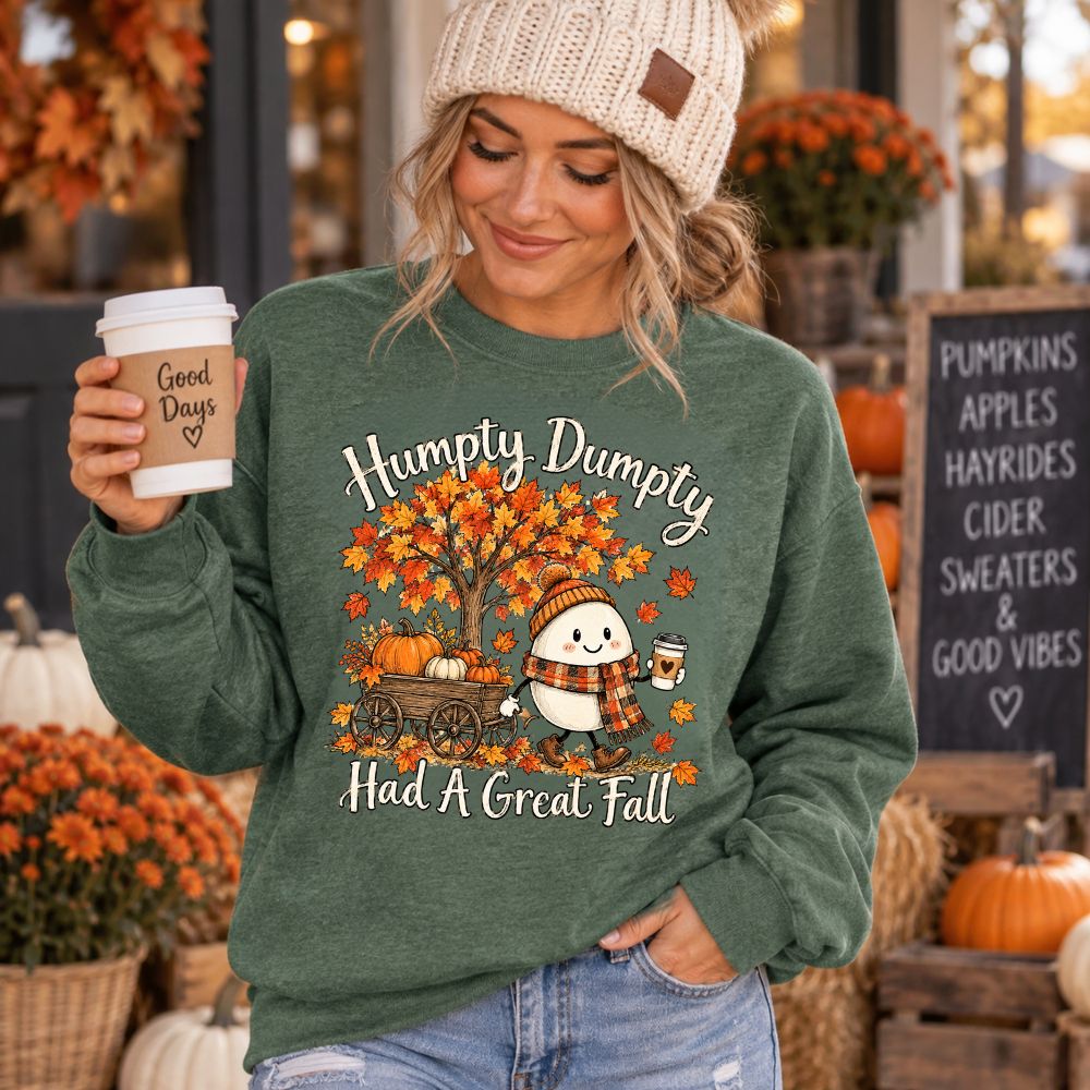 A woman wearing a forest green crewneck sweatshirt with a fall-themed graphic that reads 'Humpty Dumpty Had A Great Fall' and shows a cartoon egg under an autumn tree holding a coffee cup and wearing a cream knit beanie in an outdoor autumn-themed setting with pumpkins and a chalkboard sign.