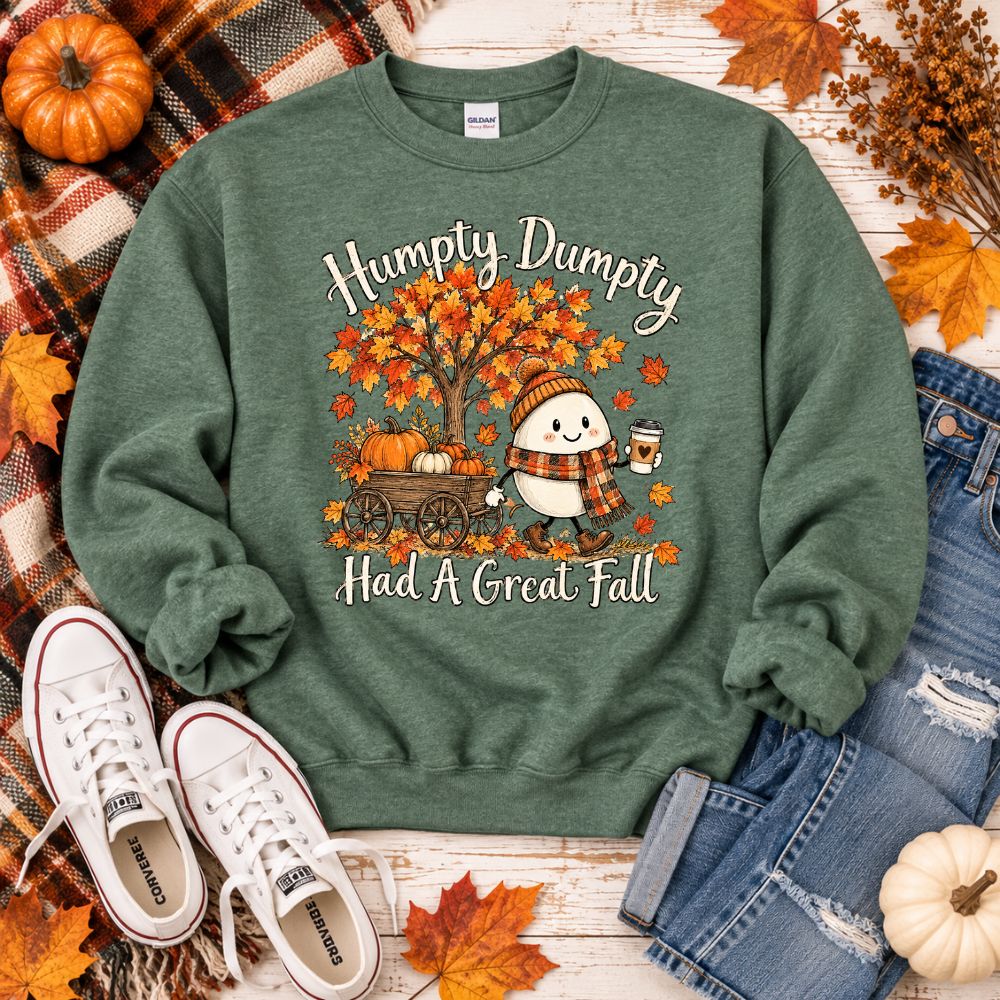 Heather green crewneck sweatshirt with "Humpty Dumpty Had a Great Fall" on the front with a egg dressed in a hat, scarf, holding a hot beverage next to a fall tree and pumpkins.  Shirt is laying on wood background with jeans, shoes, and plaid scarf. 