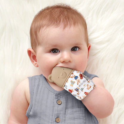 Baby wearing and using the tan teddy bear teething mitt laying on a white fur background.