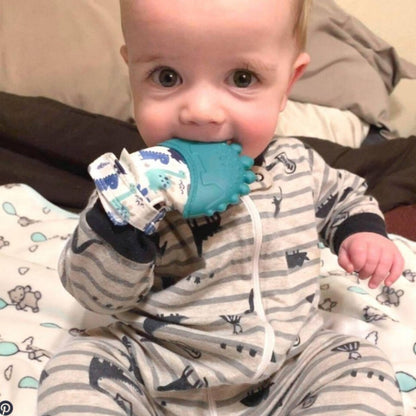 Baby wearing and chewing on Green Dinosaur Teething Mitt sitting on bed.