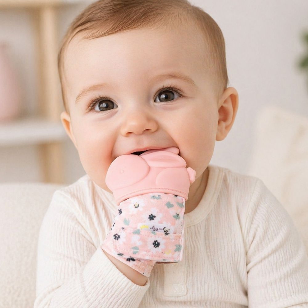 Baby wearing pink bunny teething mitt chewing on it on a white background.