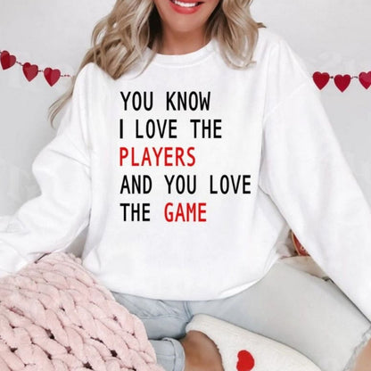KC Love the Players Love the Game Sweatshirt. White fleece lined crewneck unisex sweatshirt with black and red play on lyrics sweatshirt on model with hearts in background on model sitting with shirt on and hearts in background. Little Gift Nook.