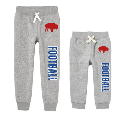 Two pairs of heather gray kids sweatpants shown side by side, both with white drawstrings, a red buffalo graphic on the upper right leg, and vertical blue 'FOOTBALL' text along the leg.