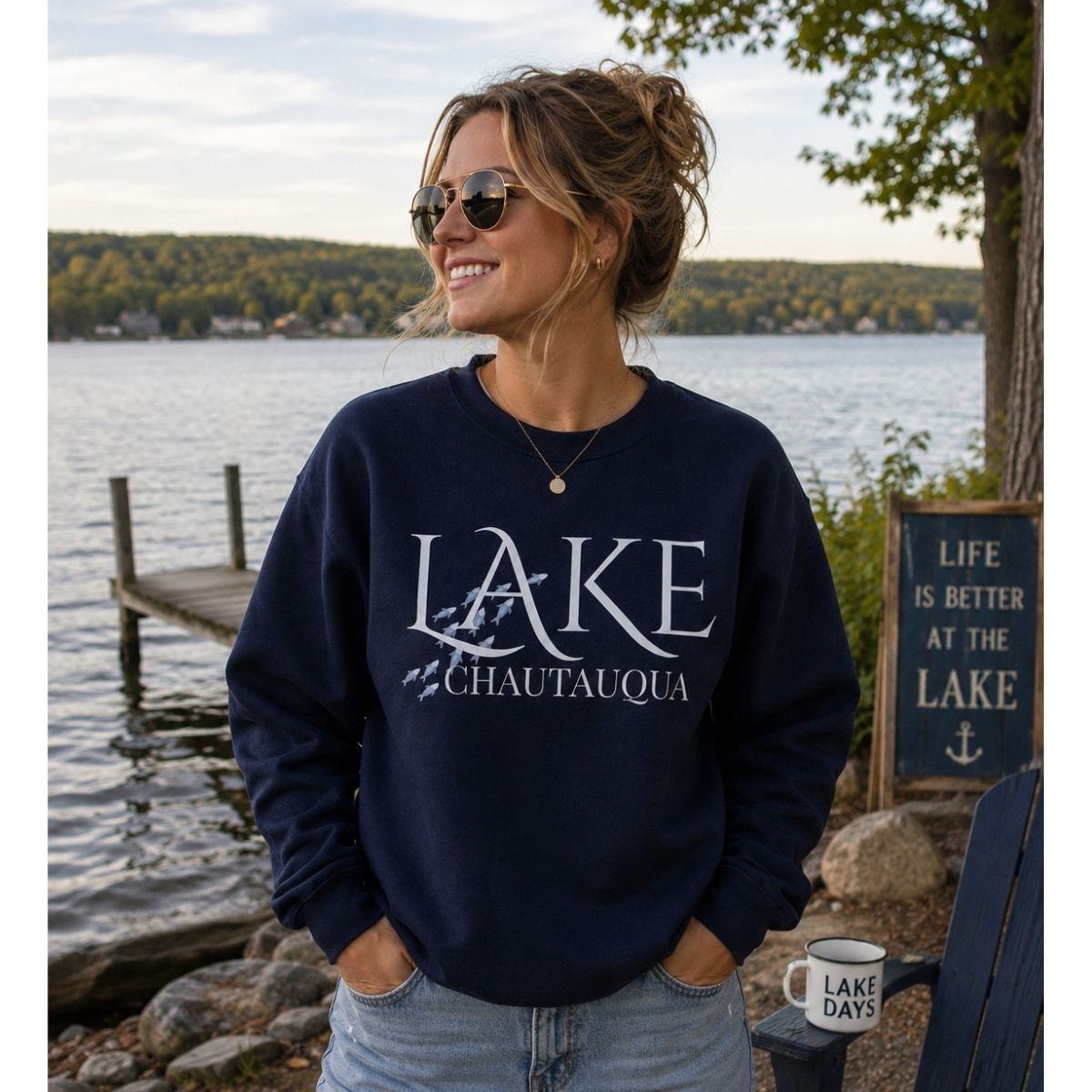 Woman wearing navy "Lake Chautauqua" sweatshirt with blue fish swimming by the word LAKE standing in front of lake and dock - Little Gift Nook