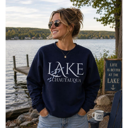 Woman wearing navy "Lake Chautauqua" sweatshirt with blue fish swimming by the word LAKE standing in front of lake and dock - Little Gift Nook