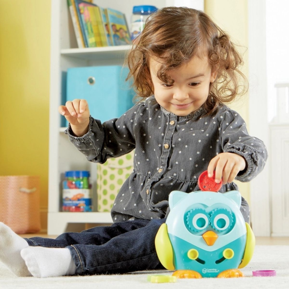 Little toddler girl playing with Learning Resources Hoot the Owl toy putting plastic dics into slit sitting on carpet. 