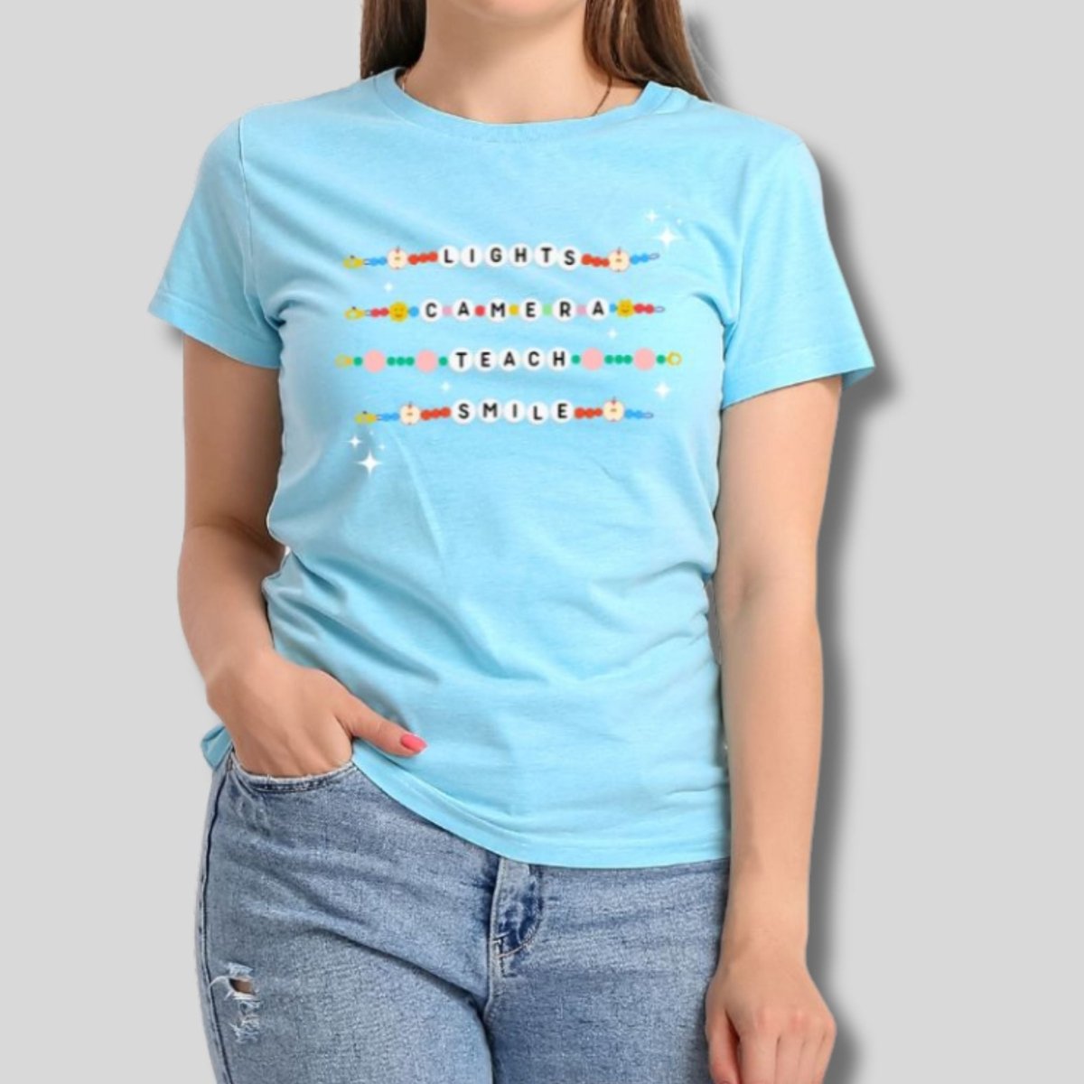 Woman wearing Light blue tshirt with friendship bracelets beads that say Lights, Camera, Teach, Smile in letter beads on front standing on light gray background.