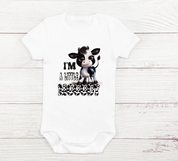 white baby onesie with black white cow and "I'm a little mooody" on front sitting on white wood background.