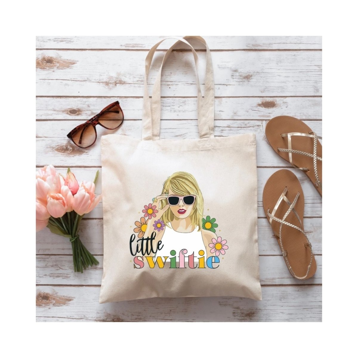 Beige tote bag with little Swiftie on front and image of singer in glasses with flowers sitting on faux wood background with flip flops and sunglasses. 