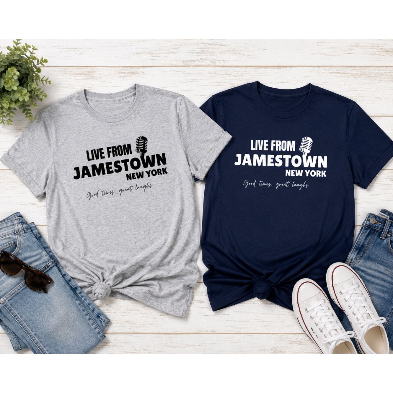 Two graphic t-shirts displayed in a flat-lay arrangement with jeans, sunglasses, and sneakers on a white wood background. One t-shirt is light heather gray with black text reading 'LIVE FROM JAMESTOWN NEW YORK' and retro microphone image. Navy shirt is the same but in white.
