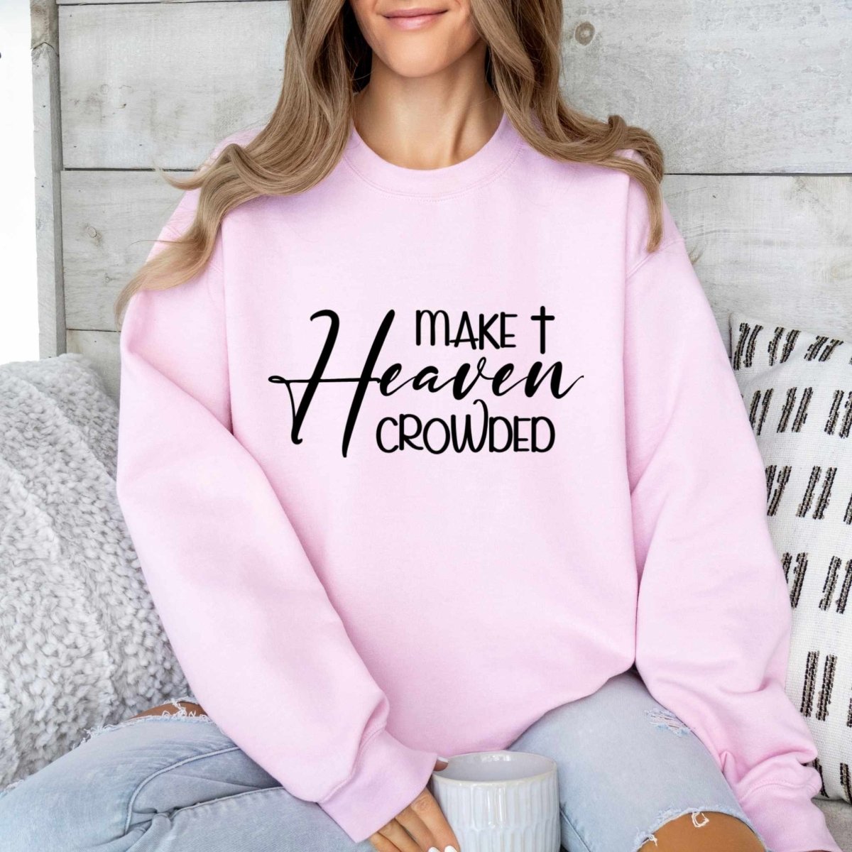 woman in pink crewneck sweatshirt with "Make Heaven Crowded" in black text on front on a sofa and gray background.