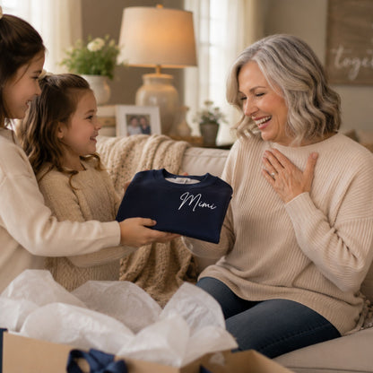 A navy blue crewneck sweatshirt with 'Mimi' written in white script is held up in a cozy living room setting. A young girl in a cream sweater holds the sweatshirt while a smiling woman with gray hair sits on a couch in the background.