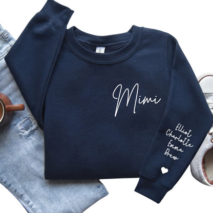A navy blue crewneck sweatshirt laid flat on a white background, styled with light-wash denim jeans, a small brown cup, and a gray shoe. The sweatshirt has 'Mimi' written in white script on the chest and a list of names on one sleeve with a small white heart.