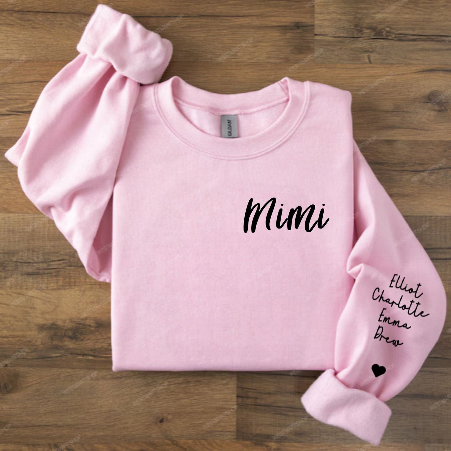 A light pink long-sleeve sweatshirt laid flat on a wooden surface, with the word 'Mimi' printed in black script across the chest and additional black kids names and a small heart printed on one sleeve.