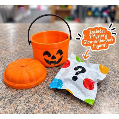 An mini orange plastic pumpkin-shaped bucket with a black handle and a matching orange lid sits on a speckled countertop. A white foil mystery pack with colorful dots and a black question mark is placed beside it, with an orange callout bubble reading 'Includes 1 Mystery Glow-in-the-Dark Figure!'.