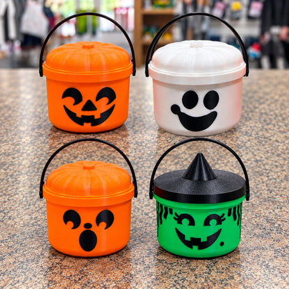 Four plastic mini Halloween buckets arranged in a 2x2 grid on a speckled countertop. The top-left bucket is orange with a pumpkin face and black handle, then white with black face as a ghost, and green and black witch hat lid on 4th.