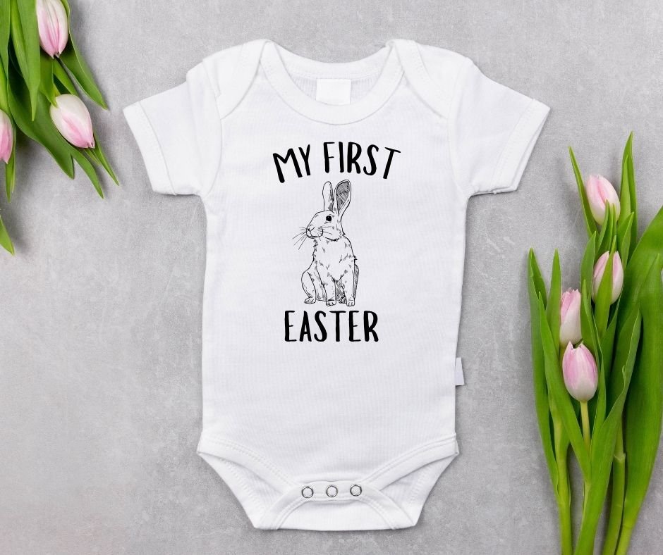 White baby bodysuit with "My First Easter" and black sketched bunny graphic on front laying on gray background with pink tulips.