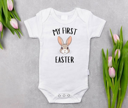 White baby bodysuit with "My First Easter" and pink bunny ear graphic on front laying on light gray background with pink tulips. 