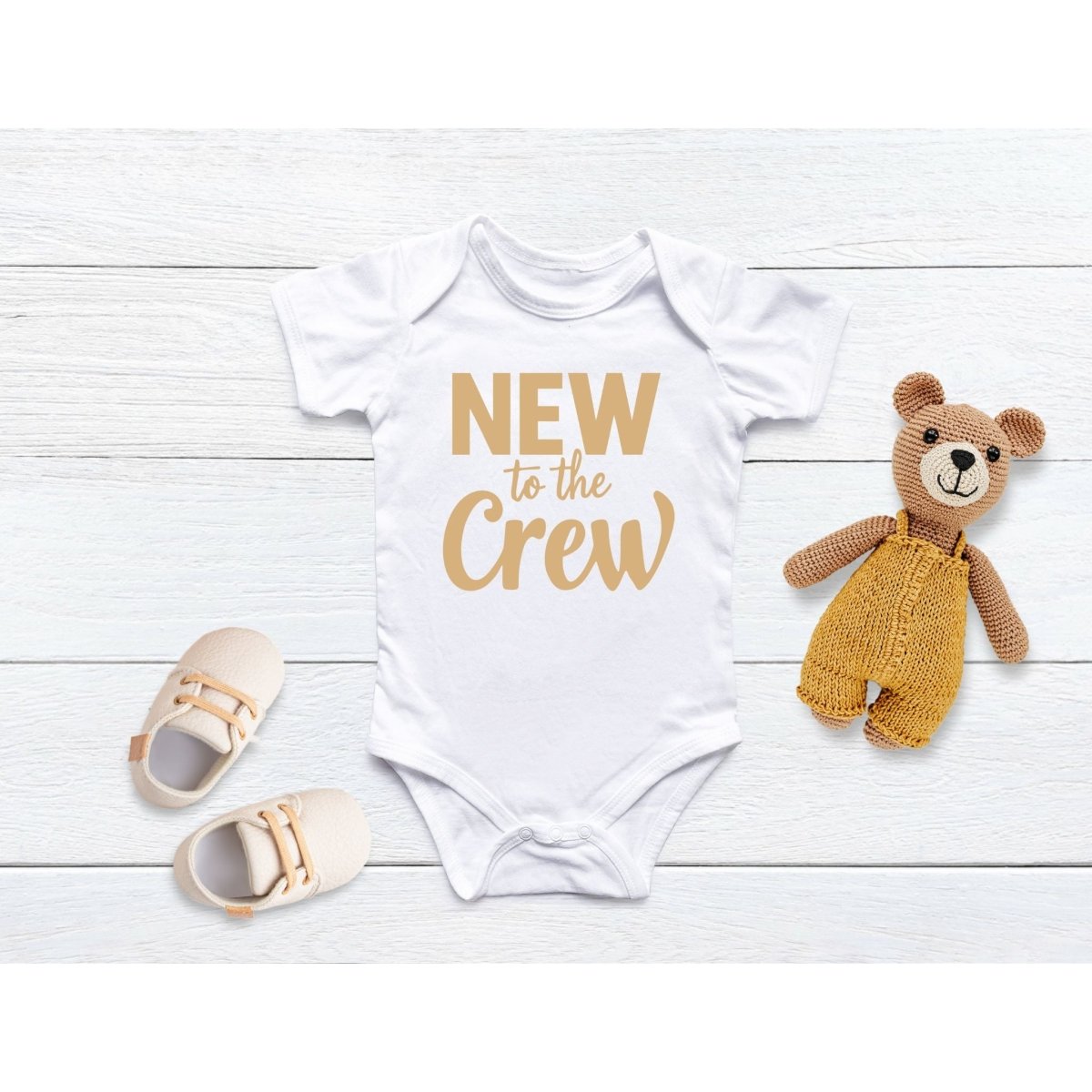 White and tan lettered baby onesie "New to the Crew" on a faux wood background with baby shoes and teddy bear. 