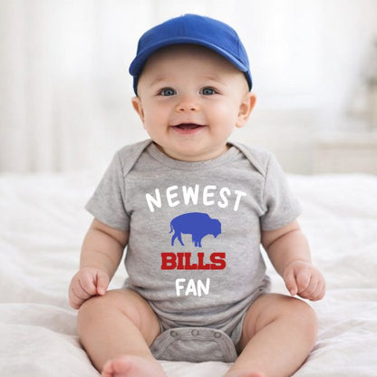 Baby wearing gray onesie with "Newest Bills fan" and blue buffalo on front in a blue baseball hat on a white blanket background.