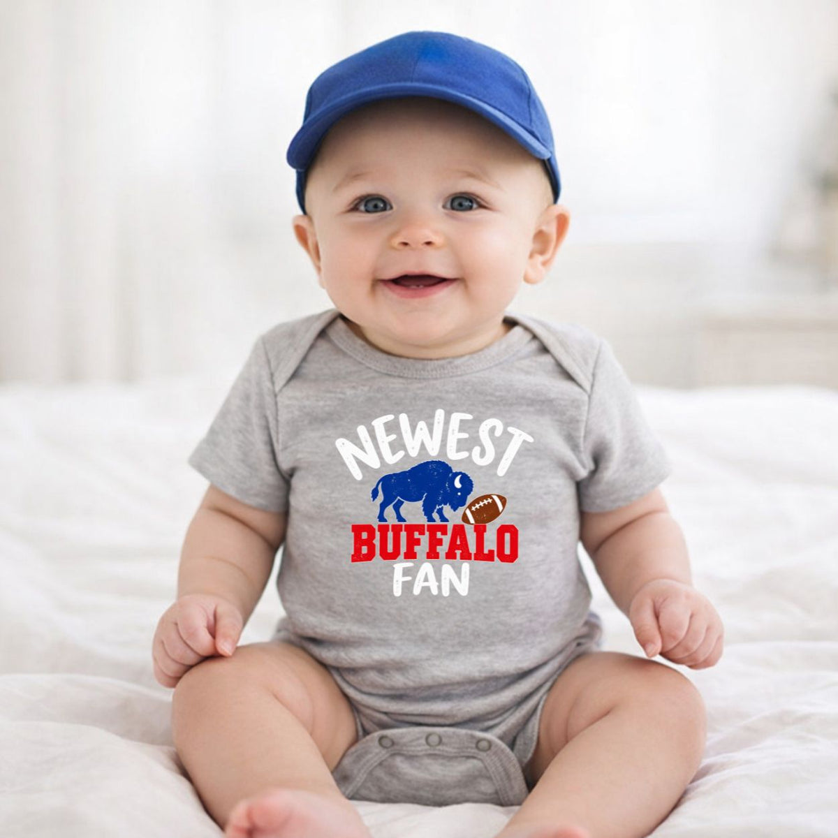 baby wearing blue baseball hat and gray bodysuit with "Newest Buffalo fan" on front and blue buffalo and brown football.