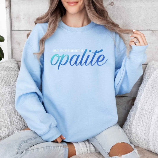Woman wearing light blue crewneck sweatshirt with white "BUT NOW THE SKY IS" AND "OPALITE" in Iridescent blue lettering on front sitting on a creme couch background. Little Gift Nook