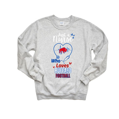 A light heather gray crewneck sweatshirt featuring a graphic design on the front. The design includes a blue heart outline with a red charging buffalo inside, a stethoscope, and the text 'Just a Nurse Who Loves BUFFALO FOOTBALL' in white, blue, and red.