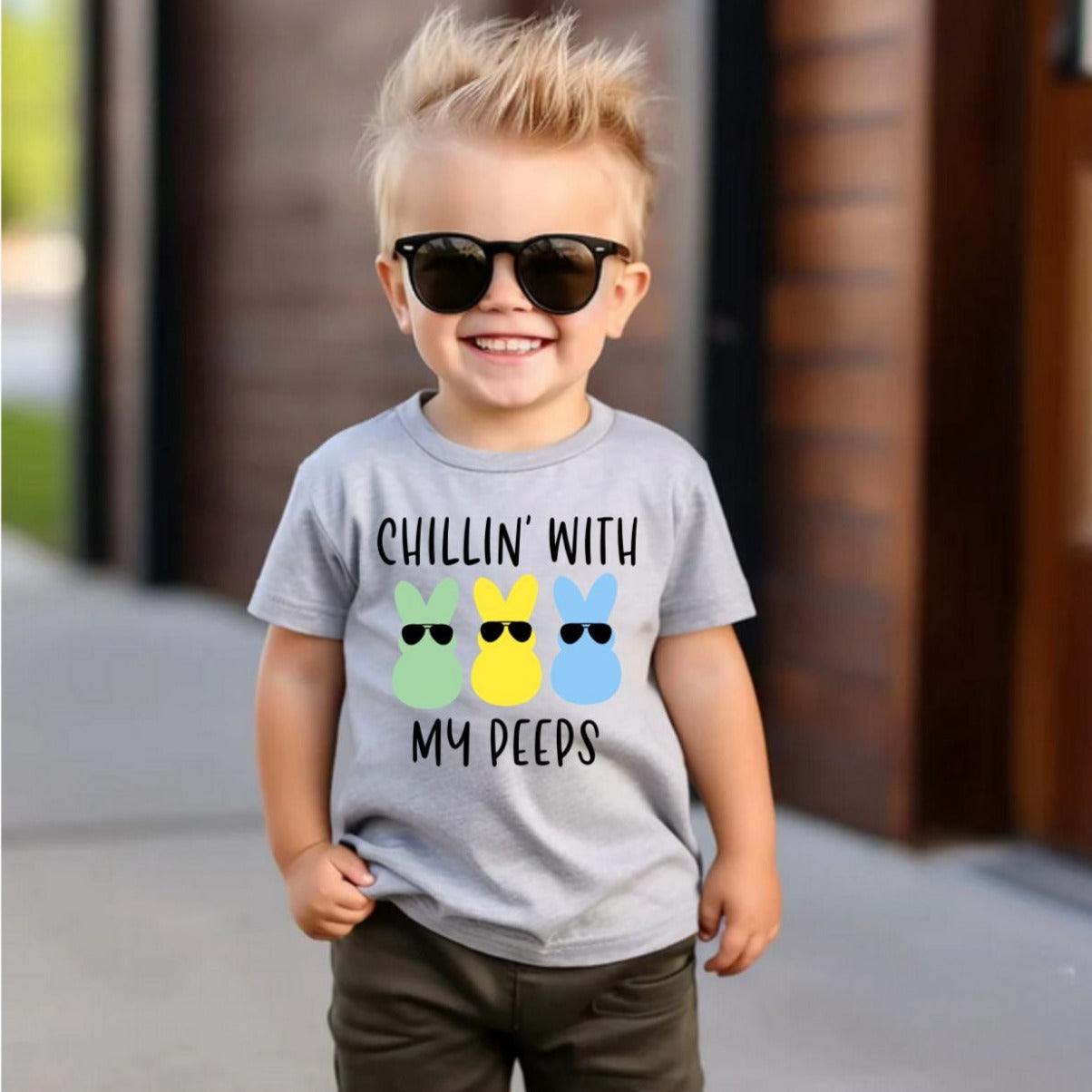 Little boy in sunglasses wearing the heather gray  Chillin' With My Peeps Kids Easter Tshirt . Black matte lettering with green, yellow, and blue bunny peep in sunglasses on front of shirt. Little Gift Nook.