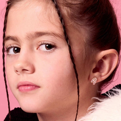 Close-up of a young girl with braided hair wearing pink ballerina slipper stud earrings on a pink background. 