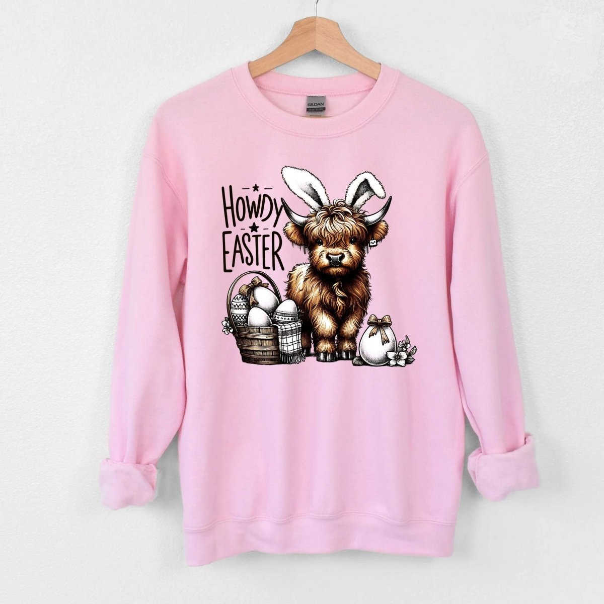 Pink sweatshirt with cow wearing bunny ears with easter eggs and basket "Howdy Easter" on front hanging on a light gray background. Little Gift Nook