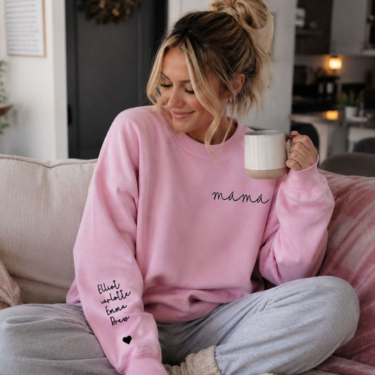 Woman wearing pink crewneck sweatshirt with "mama" in cursive on left chest and kids names on sleeve with heart, holding a cup of coffee on a couch. 