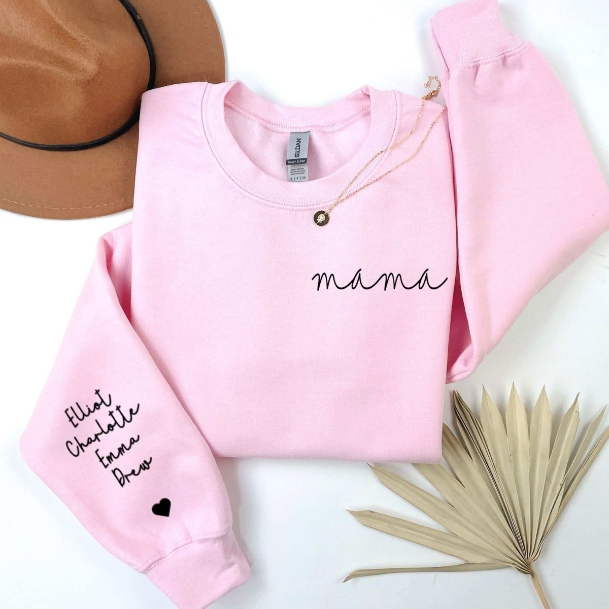 Pink crewneck sweatshirt with mama on top left and names on sleeve on a white background with a fan and hat. Little Gift Nook