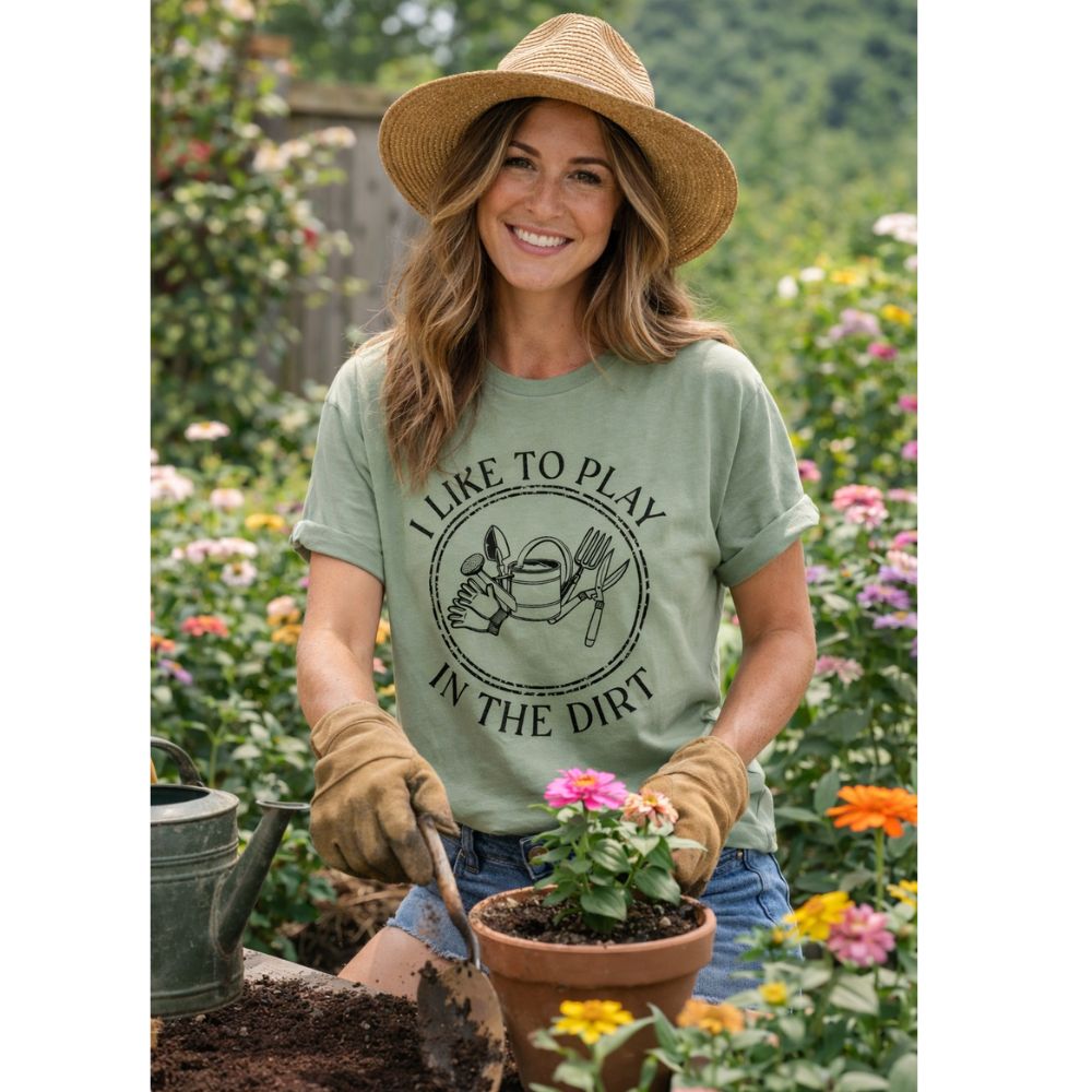 Woman wearing sage green "I Like to Play in the Dirt" tshirt with gardening tools image on front in a garden outdoors.