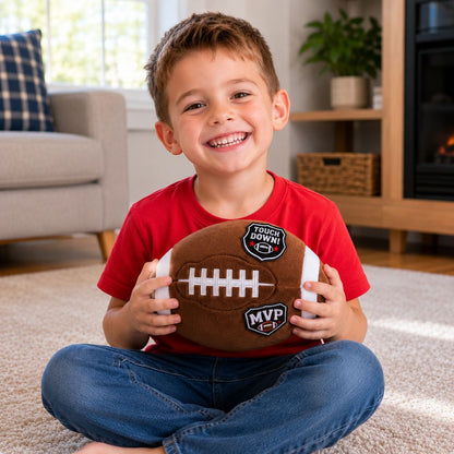 Little boy holding plush football with iron on patches on it sitting in living room. 