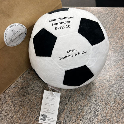 Personalized black and white plush soccer ball with baby name, birth date, and message on colorful counter top with bag.