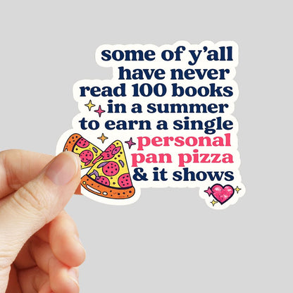 A hand holds a die-cut sticker with a humorous quote "some of ya'll have never read 100 books in summer to earn a single personal pan pizza and it shows" featuring navy blue and pink text with a cartoon pizza slice illustration on a light gray background.