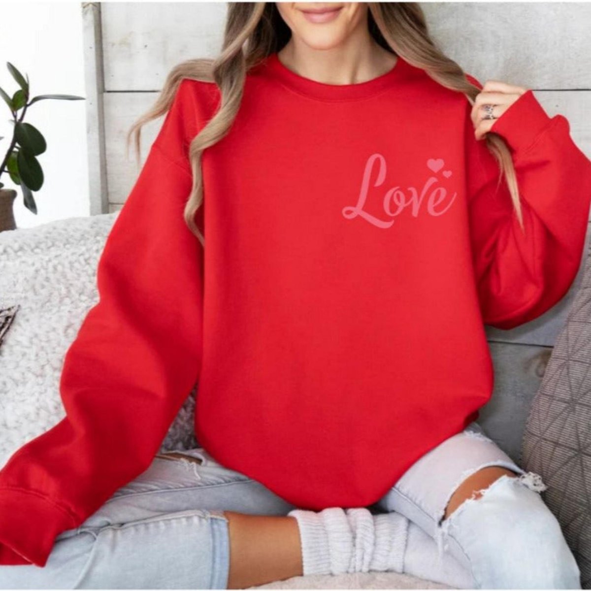 Woman wearing red crewneck sweatshirt with pink LOVE and hearts at top right chest sitting on couch. 
