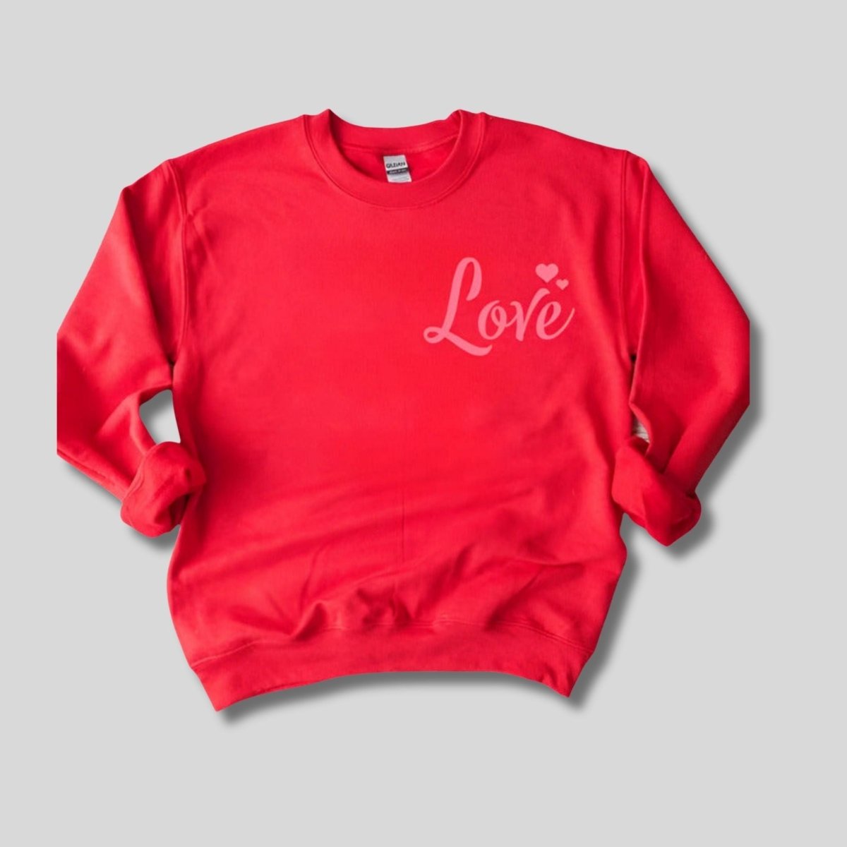 Red crewneck sweatshirt with pink LOVE and hearts at right chest sitting on gray background. Little Gift Nook