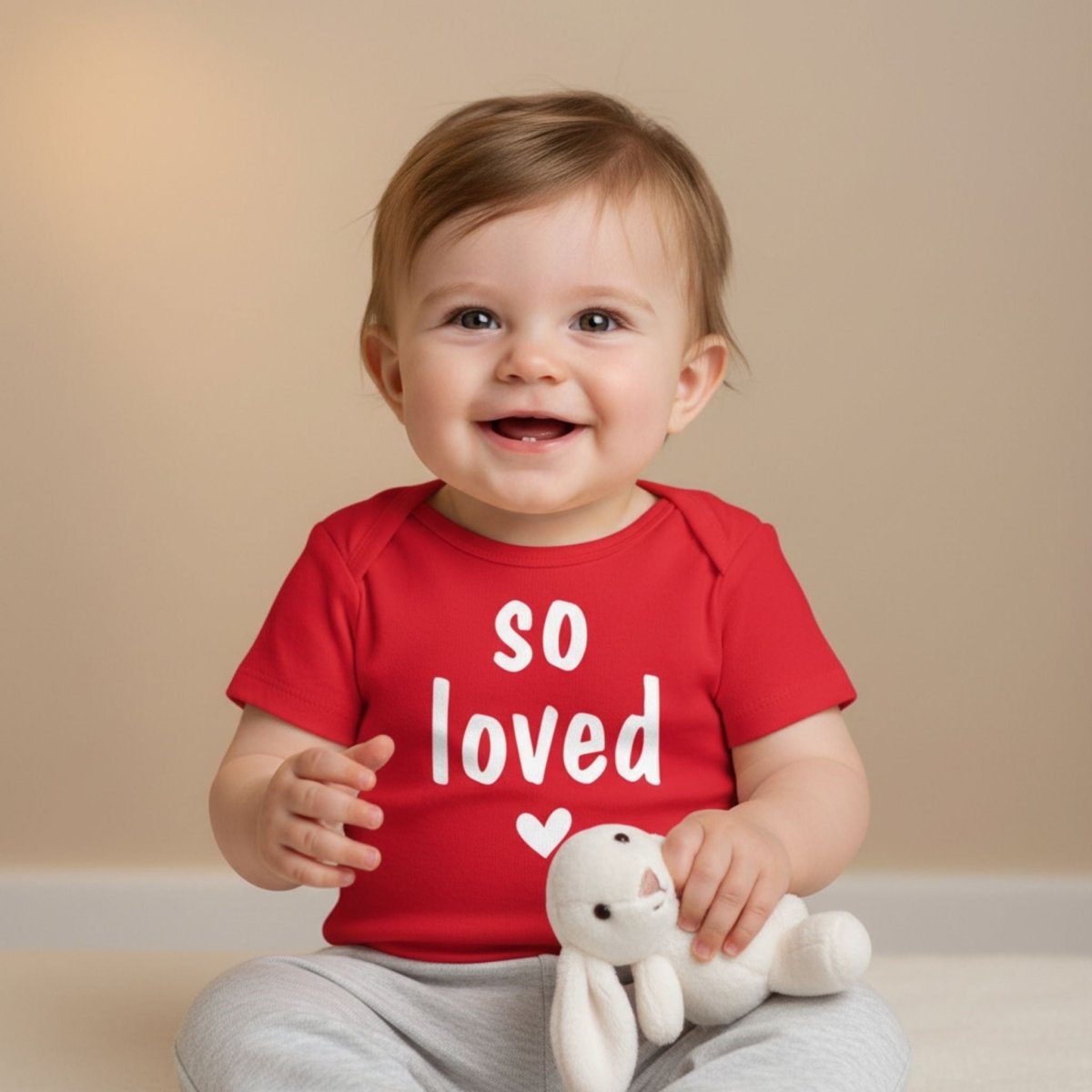 Baby wearing a red 'so loved' bodysuit onesie holding a plush toy against a beige background
