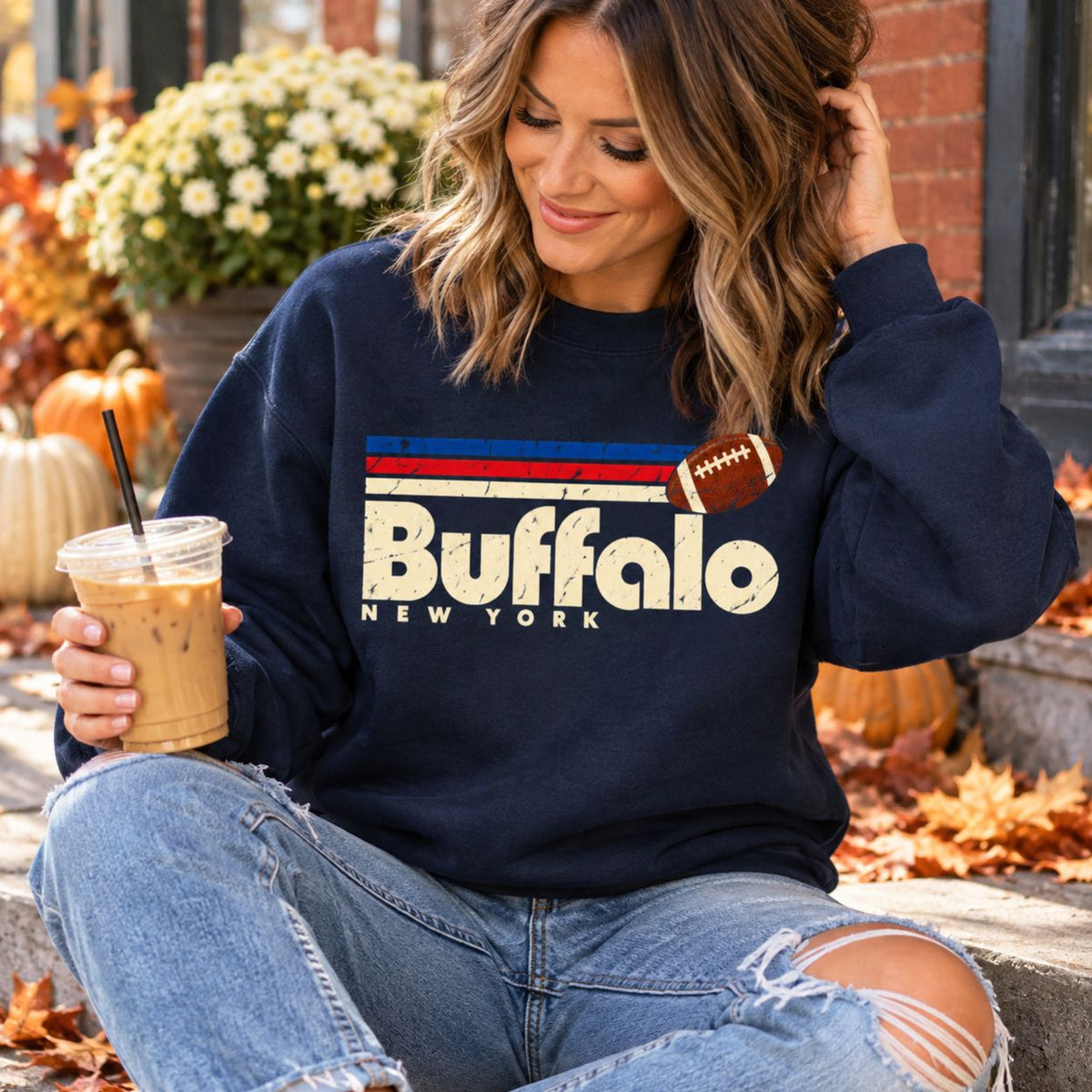 A woman sitting outdoors wearing a navy blue sweatshirt with a distressed Buffalo New York football graphic, paired with ripped blue jeans and holding an iced coffee.