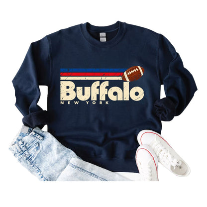 A navy blue crewneck sweatshirt with a distressed Buffalo New York graphic, styled in a flat lay with light-wash jeans and white sneakers on a white background.