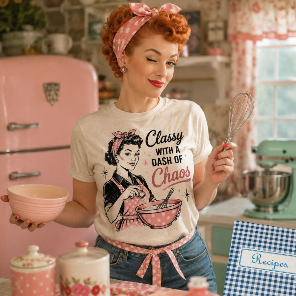 Woman wearing beige tshirt with "Classy with a Dash of Choas and woman mixing bowl in apron drawing on front standing in front of a vintage looking kitchen. 