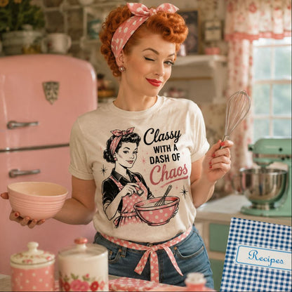 Woman wearing beige tshirt with "Classy with a Dash of Choas and woman mixing bowl in apron drawing on front standing in front of a vintage looking kitchen. 