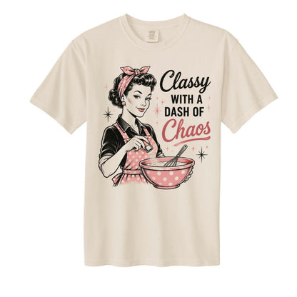 Beige woman's tshirt with image of woman in pink polkadot apron and hair scarf mixing bowl and sprinkling in ingredients and caption "Classy with a dash of Choas" on front sitting on white background.