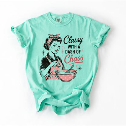 Mint green t-shirt with woman in polkadot apron and hair scarf mixing a bowl with text " Classy with a dash of Chaos" on front sitting on white background. 