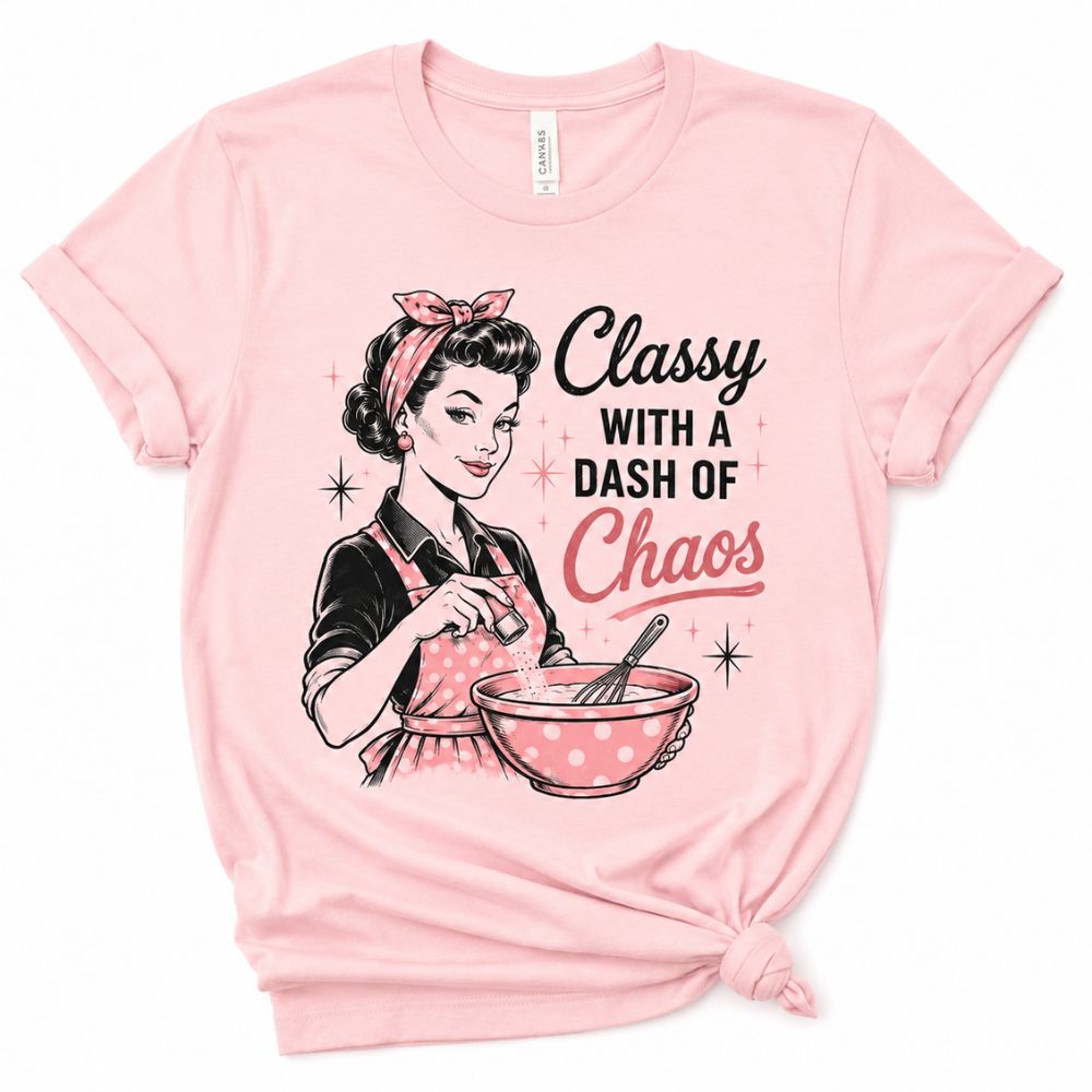 Pink t-shirt with image of woman in apron mixing a bowl and text "Classy with a Dash of Chaos" on front sitting on white background.
