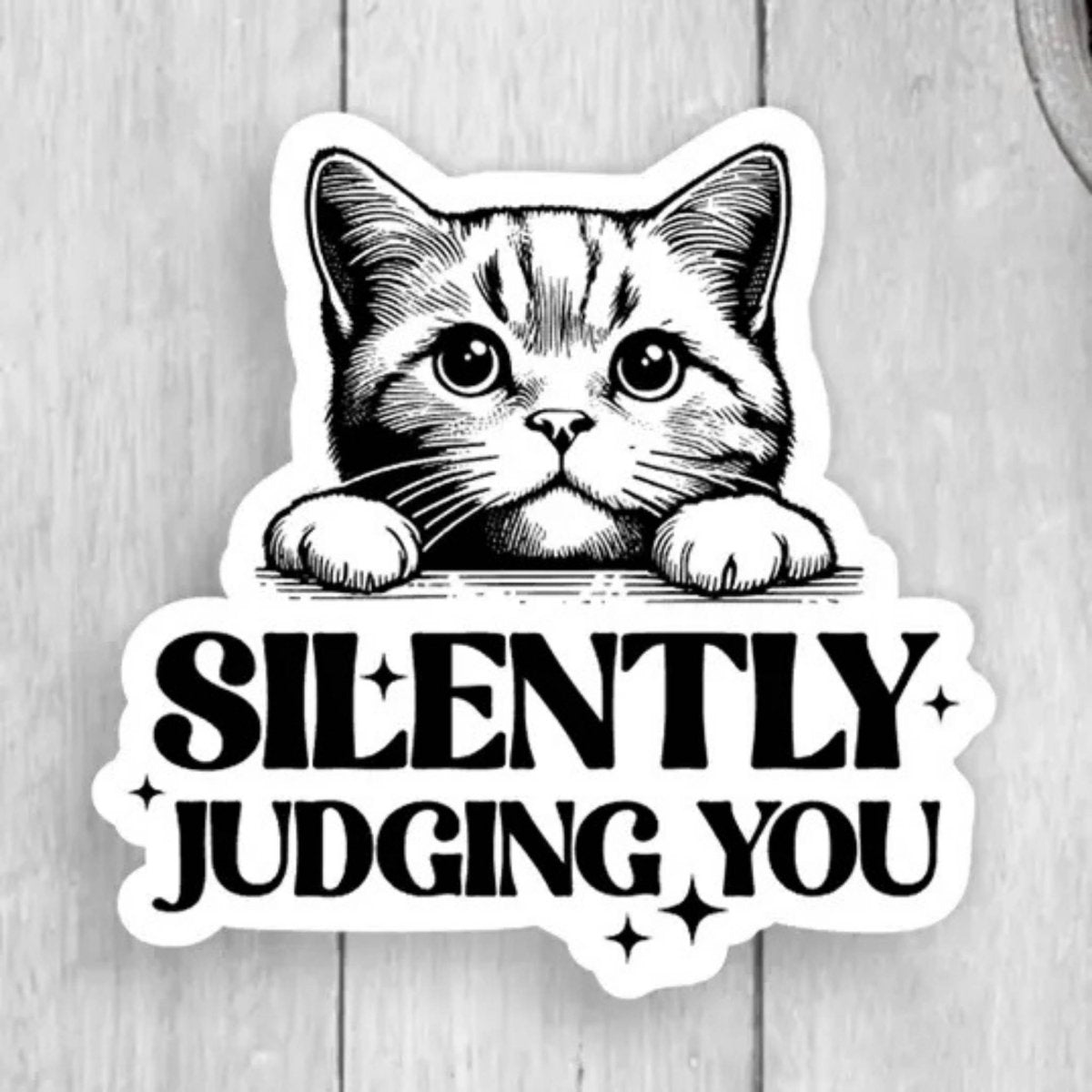 Sticker with cat peaking over text "Silently Judging You" black and white sticker on wood background. 